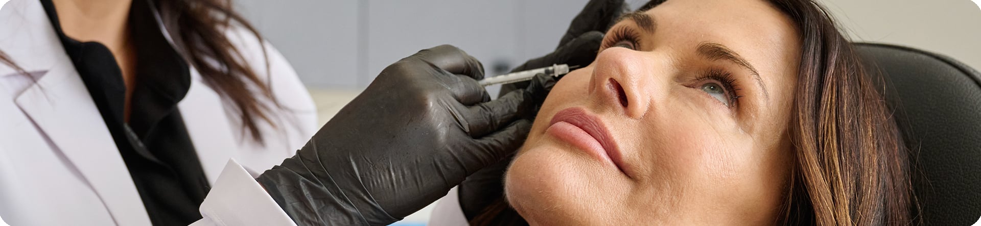 Image: Close-up photograph capturing a woman receiving a facial injectable treatment. A healthcare professional, dressed in a white coat and black gloves, carefully administers a needle near the woman's eye area. The focus is on the precise procedure, emphasizing professionalism and care in aesthetic treatments.