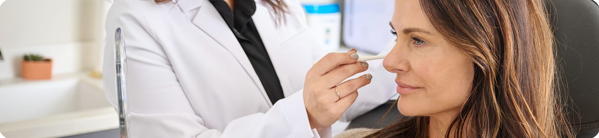 Image: Close-up photograph of a woman undergoing a consultation for injectable treatment. A healthcare professional, holding a pointer, gestures towards specific areas on the client's face while discussing the treatment plan. The focus is on the interaction between the client and the professional, highlighting the informative and personalized nature of the consultation process.